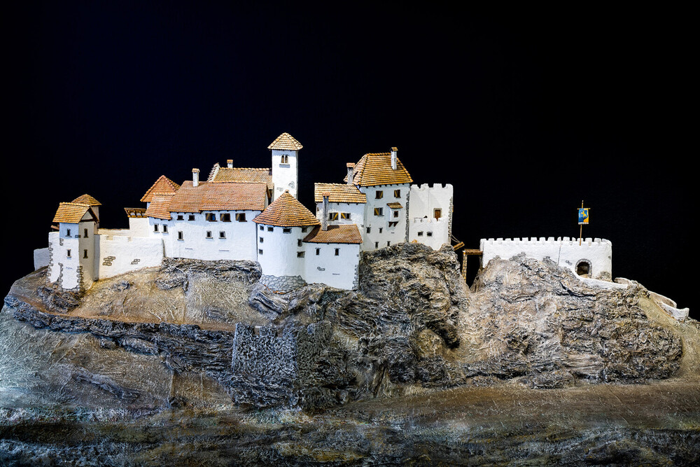 Eine detaillierte Modellansicht einer weißen Burg mit mehreren Türmen und roten Dächern, die auf einem Felsen steht. Der Hintergrund ist schwarz.