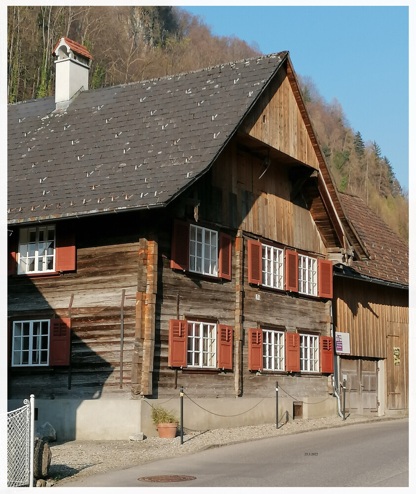 Ein Holzhaus mit einem steilen Schindeldach, das neben einer Straße steht. Im Hintergrund befindet sich ein bewaldeter Hang unter einem klaren Himmel.